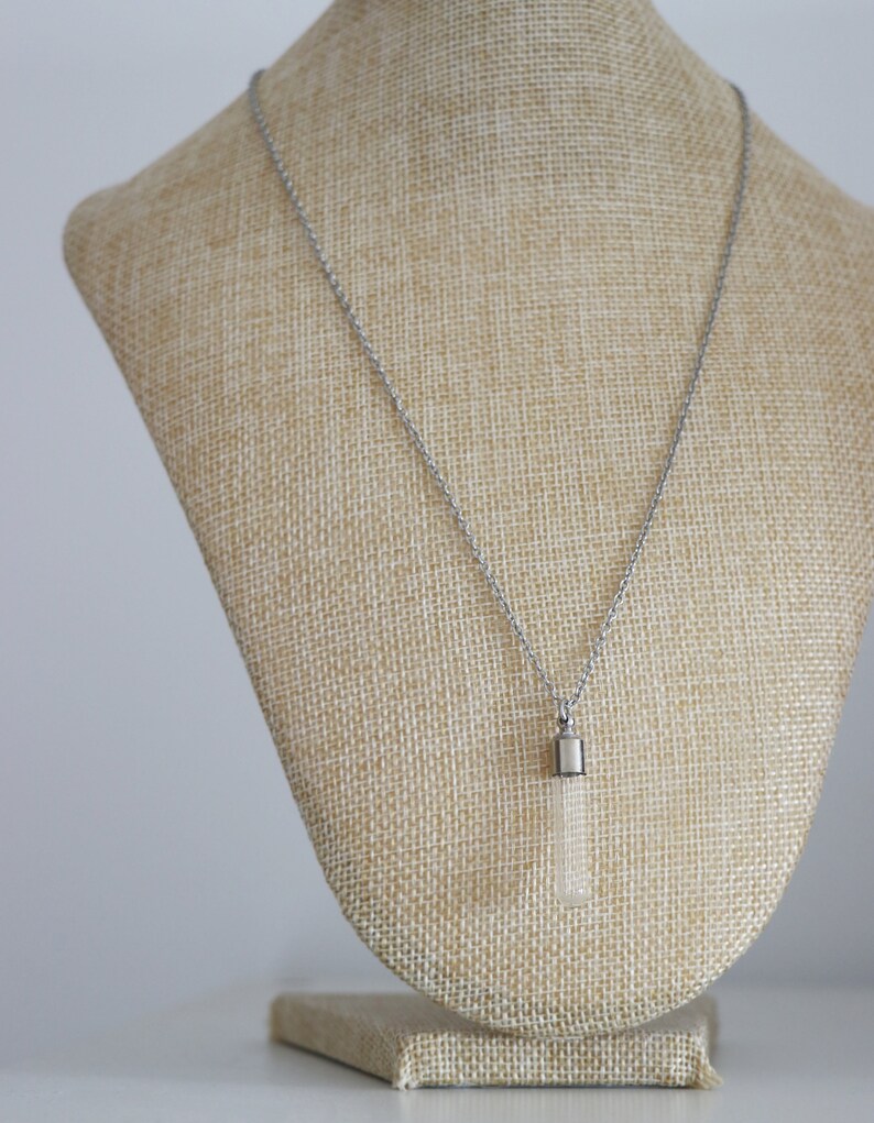 May include: A silver necklace with a rectangular, clear pendant. The pendant is suspended from a delicate chain and is displayed on a beige bust. The necklace is a simple, elegant piece of jewelry.