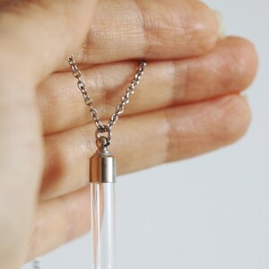 May include: A clear glass vial pendant necklace with a silver-colored chain. The vial has a silver-colored cap and is held in a hand. The necklace is a simple, elegant accessory.