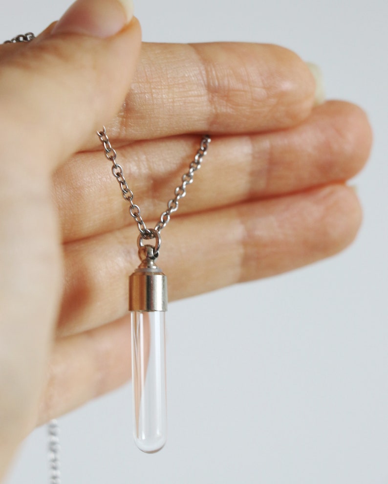 May include: A clear glass vial pendant necklace with a silver-colored chain. The vial has a silver-colored cap and is held in a hand. The necklace is a simple, elegant accessory.