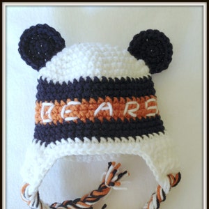 Peut inclure: Bonnet b&eacute;b&eacute; en crochet blanc avec des rayures noires et orange et le mot "BEARS" brod&eacute; sur le devant. Le bonnet a deux oreilles en crochet noir.