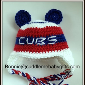 May include: A red, white, and blue crocheted baby hat with ear flaps and a braided tie. The hat has the word "CUBS" embroidered on the front.
