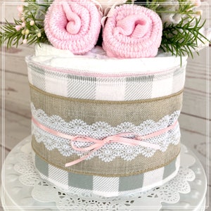 May include: A diaper cake decorated with a gray and white plaid fabric, a burlap ribbon, and a white lace ribbon with a pink bow. The cake is topped with two pink baby socks and a sprig of greenery.