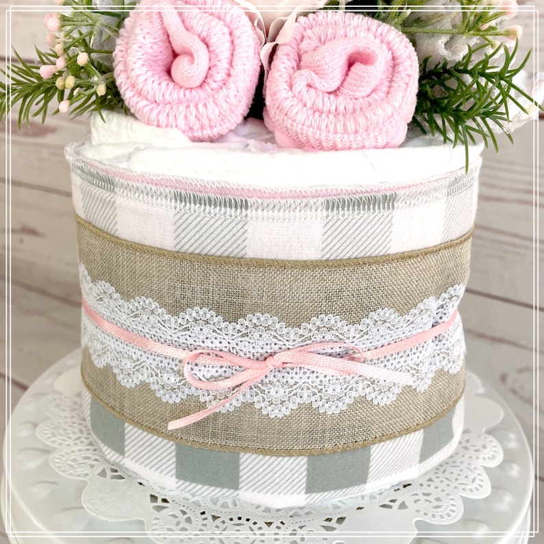 May include: A diaper cake decorated with a gray and white plaid fabric, a burlap ribbon, and a white lace ribbon with a pink bow. The cake is topped with two pink baby socks and a sprig of greenery.