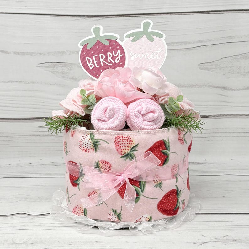 May include: A pink and white diaper cake decorated with pink flowers, green leaves, and two strawberry-shaped signs that say "Berry Sweet".