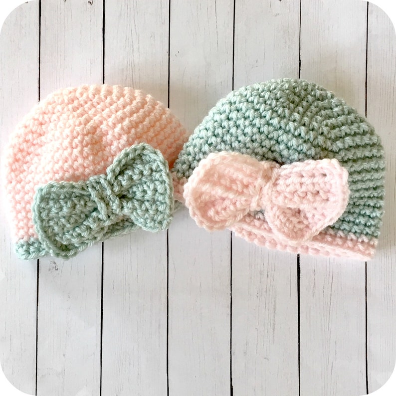 Peut inclure: Deux bonnets pour b&eacute;b&eacute; en crochet avec des n&oelig;uds. Un bonnet est rose avec un n&oelig;ud vert, et l'autre est vert avec un n&oelig;ud rose.