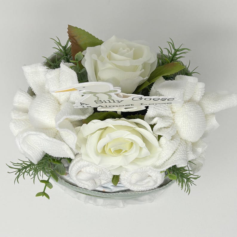 May include: A white floral arrangement featuring white roses and towel creations, accented with green foliage. A small sign reads "A Silly Goose Is Almost Loose."