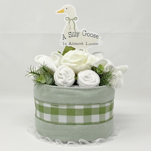 May include: A baby gift basket with a light green exterior and a green and white checkered ribbon. The basket is decorated with white flowers, greenery, and rolled-up baby items. A sign with a goose illustration and the text "A Silly Goose Is Almost Loose" is on top.