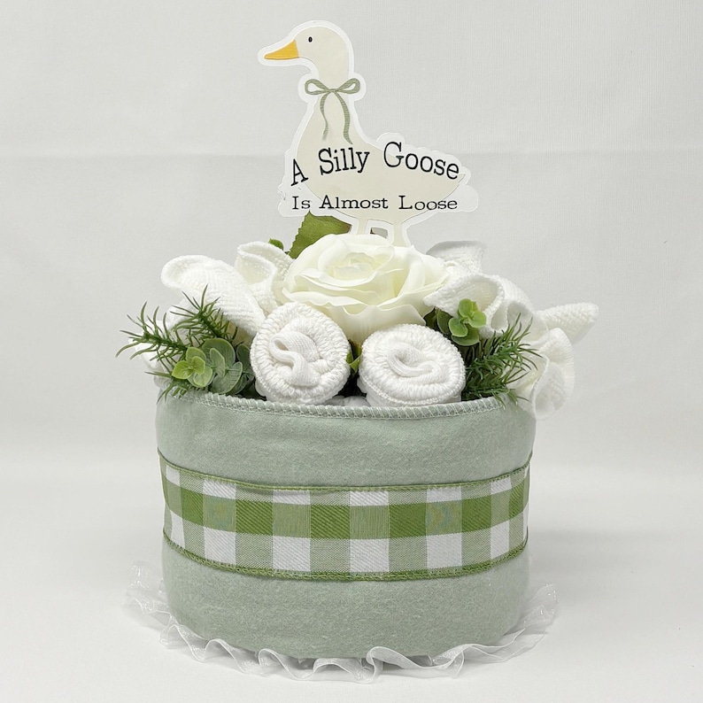 May include: A baby gift basket with a light green exterior and a green and white checkered ribbon. The basket is decorated with white flowers, greenery, and rolled-up baby items. A sign with a goose illustration and the text "A Silly Goose Is Almost Loose" is on top.