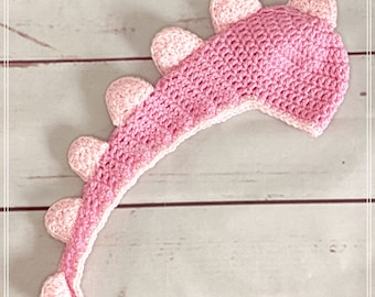 May include: Pink crocheted dinosaur tail with a pointed tip and a row of pink crocheted spikes along the top.