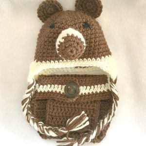 Peut inclure: Chapeau b&eacute;b&eacute; en crochet marron avec des oreilles d'ours et un bouton marron. Le chapeau a une bordure en crochet blanc et une cravate tress&eacute;e blanche et marron.