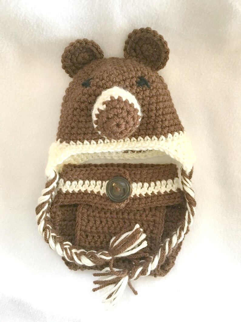 Peut inclure: Chapeau b&eacute;b&eacute; en crochet marron avec des oreilles d'ours et un bouton marron. Le chapeau a une bordure en crochet blanc et une cravate tress&eacute;e blanche et marron.