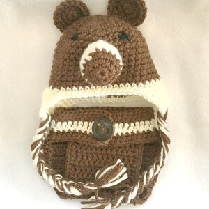 Peut inclure: Chapeau b&eacute;b&eacute; en crochet marron avec des oreilles d'ours et un bouton marron. Le chapeau a une bordure en crochet blanc et une cravate tress&eacute;e blanche et marron.