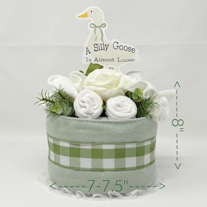May include: A light green baby gift basket, approximately 8 inches tall and 7-7.5 inches wide, decorated with white flowers, rolled baby items, and a green and white gingham ribbon. A sign with a goose graphic reads "A Silly Goose Is Almost Loose."