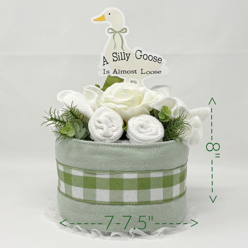 May include: A light green baby gift basket, approximately 8 inches tall and 7-7.5 inches wide, decorated with white flowers, rolled baby items, and a green and white gingham ribbon. A sign with a goose graphic reads "A Silly Goose Is Almost Loose."