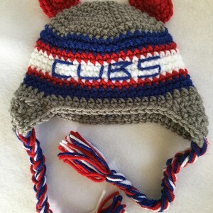 May include: Gray, blue, red, and white crocheted hat with ear flaps and a braided tassel. The hat has the word "CUBS" embroidered on the front.