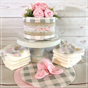 May include: A baby shower gift basket with a diaper cake decorated with pink and gray gingham, a pink bow, and a lace ribbon. The basket also includes pink socks, a stack of diapers, and a pink and gray gingham fabric.