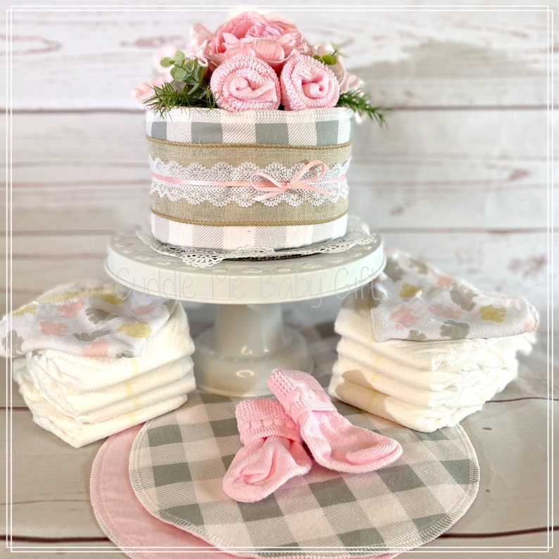 May include: A baby shower gift basket with a diaper cake decorated with pink and gray gingham, a pink bow, and a lace ribbon. The basket also includes pink socks, a stack of diapers, and a pink and gray gingham fabric.