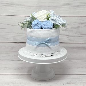May include: A white diaper cake decorated with blue and white striped fabric, blue rolled washcloths, and a white flower. The cake is on a white cake stand.