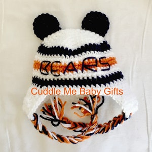 Peut inclure: Bonnet b&eacute;b&eacute; en crochet blanc, orange et bleu marine avec des pompons sur le dessus et un pompon tress&eacute;. Le bonnet est brod&eacute; du mot "BEARS" sur le devant.