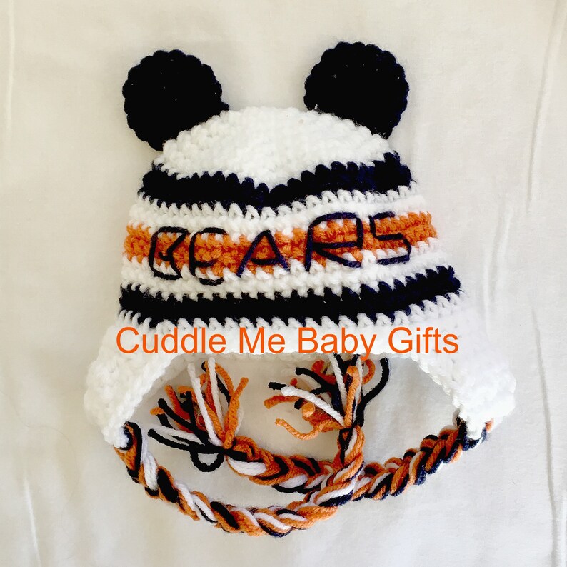 Peut inclure: Bonnet b&eacute;b&eacute; en crochet blanc, orange et bleu marine avec des pompons sur le dessus et un pompon tress&eacute;. Le bonnet est brod&eacute; du mot "BEARS" sur le devant.