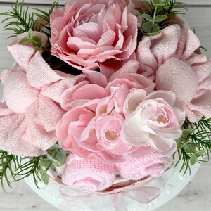 May include: A pink and white flower arrangement with pink towels and baby socks. The arrangement is in a white basket and is decorated with greenery.