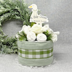 May include: A light green baby gift basket with a white and green gingham ribbon. The basket is decorated with white roses, rolled washcloths, and greenery. A sign with a cartoon goose and the text "A Silly Goose Is Almost Loose" is on top.