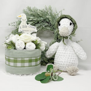 May include: A baby shower gift set featuring a white crocheted goose wearing a green bonnet, a decorative floral arrangement with a sign that reads "A Silly Goose Is Almost Loose", and a pacifier.