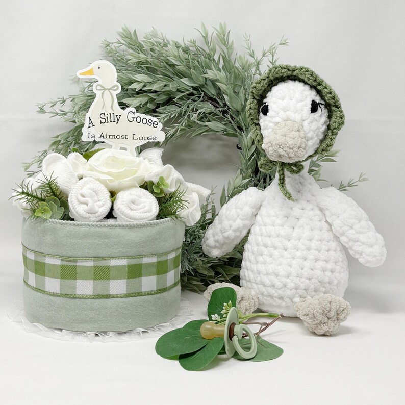 May include: A baby shower gift set featuring a white crocheted goose wearing a green bonnet, a decorative floral arrangement with a sign that reads "A Silly Goose Is Almost Loose", and a pacifier.