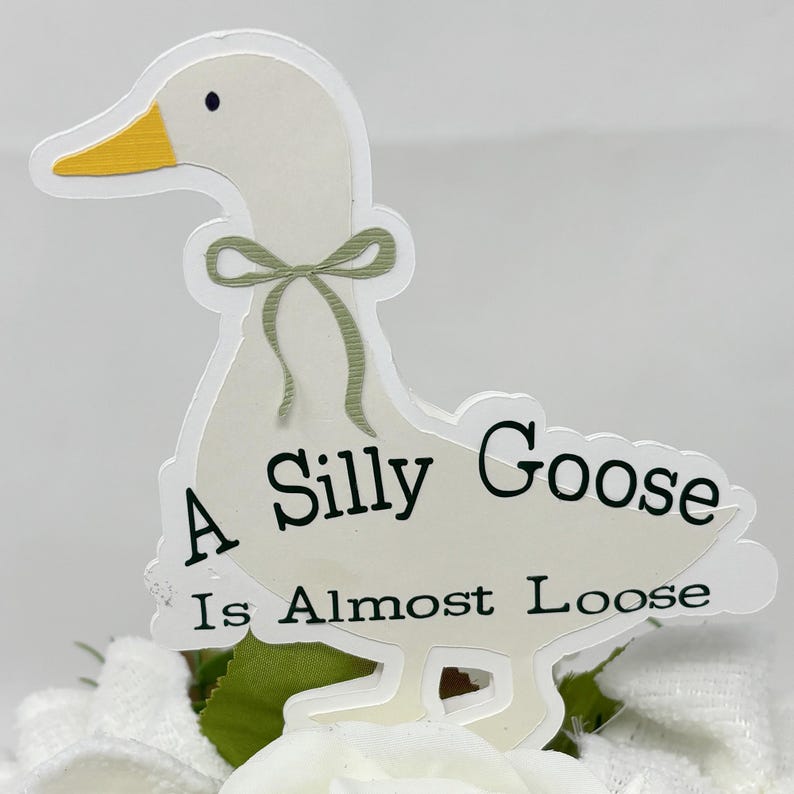 May include: A white goose-shaped sticker with a yellow beak and a green bow. The text "A Silly Goose Is Almost Loose" is printed on the goose. The sticker is placed on a white and green floral arrangement.