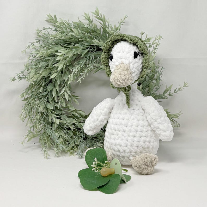 May include: A handmade white crochet goose wearing a green bonnet, with a beige beak and feet. A green wreath and a green shamrock with a pacifier are in the foreground. The goose is a soft, plush toy.
