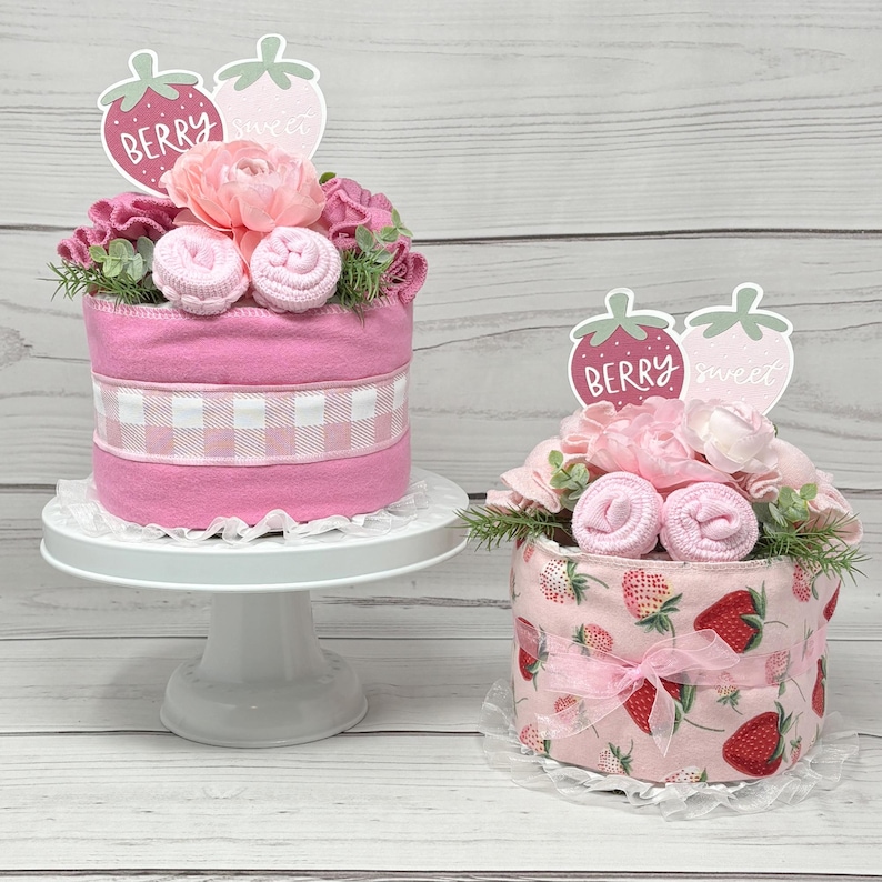 May include: Two pink and white diaper cakes decorated with pink flowers, green leaves, and strawberry-themed toppers. The toppers say "Berry Sweet".