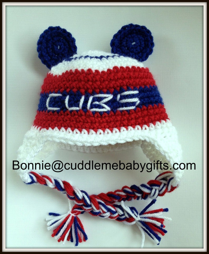 May include: A red, white, and blue crocheted baby hat with ear flaps and a braided tie. The hat has the word "CUBS" embroidered on the front.