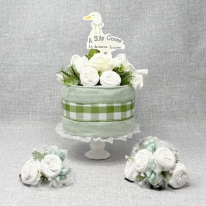 May include: A decorative baby shower cake with a light green and white gingham ribbon. The cake is topped with white flowers, greenery, and a sign that reads "A Silly Goose Is Almost Loose". Two smaller floral arrangements are also included.