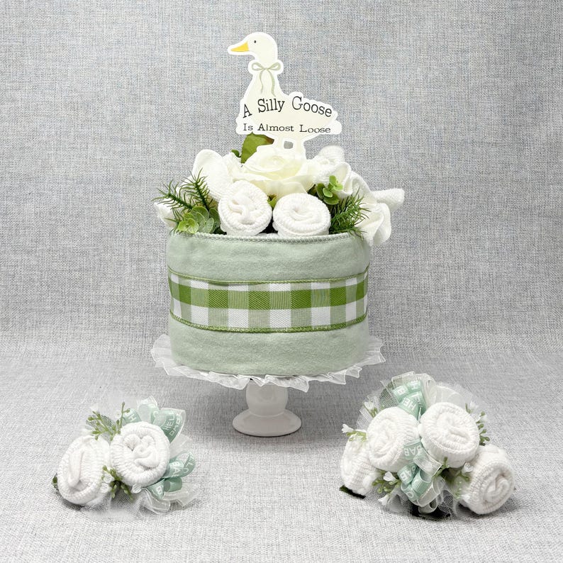 May include: A decorative baby shower cake with a light green and white gingham ribbon. The cake is topped with white flowers, greenery, and a sign that reads "A Silly Goose Is Almost Loose". Two smaller floral arrangements are also included.