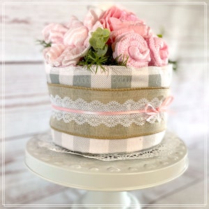 May include: A baby shower gift basket with a pink and gray checkered pattern, a burlap ribbon, and a pink lace ribbon. The basket is filled with pink and white baby items, including a pink flower.