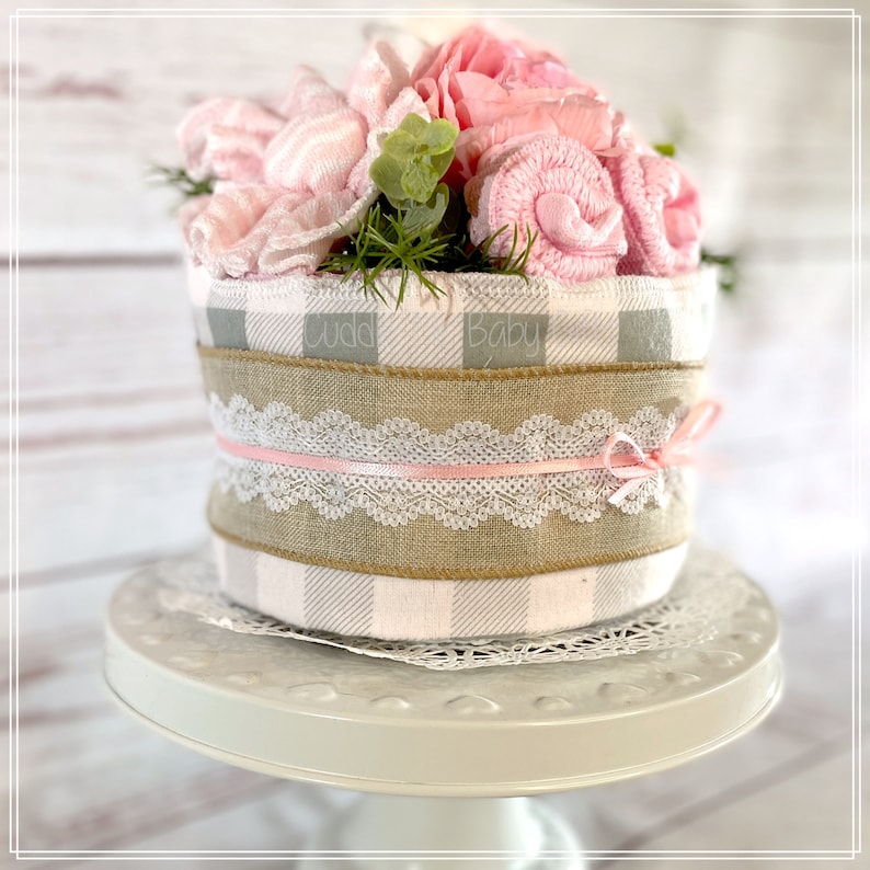 May include: A baby shower gift basket with a pink and gray checkered pattern, a burlap ribbon, and a pink lace ribbon. The basket is filled with pink and white baby items, including a pink flower.