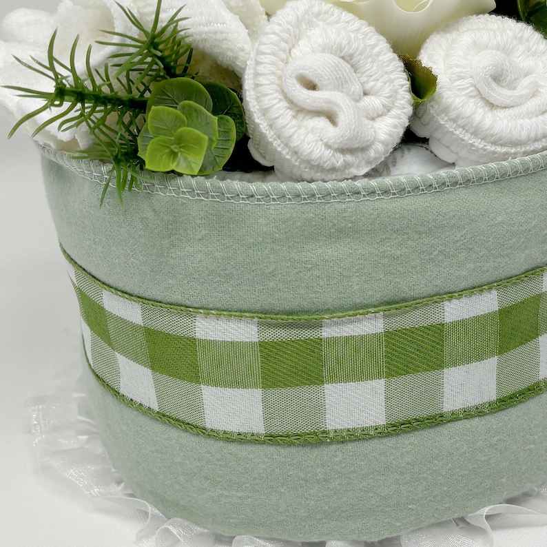 May include: A light green container with a white and green checkered ribbon. The container is filled with white rolled towels and greenery. The container has a white lace trim at the bottom.