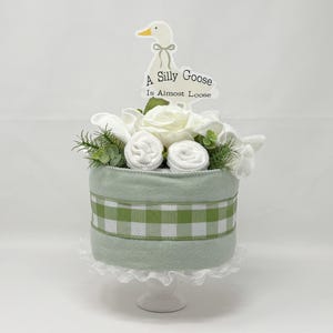 May include: A decorative baby shower cake with a light green base and a green and white gingham ribbon. The cake is topped with white flowers, rolled washcloths, and greenery. A sign with a goose illustration reads "A Silly Goose Is Almost Loose."
