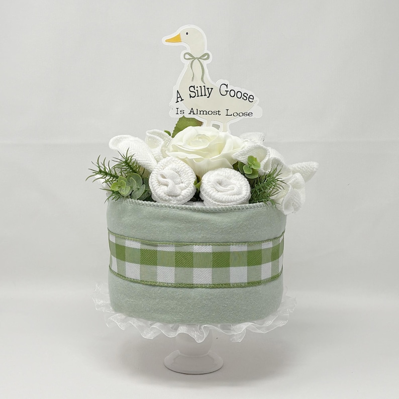 May include: A decorative baby shower cake with a light green base and a green and white gingham ribbon. The cake is topped with white flowers, rolled washcloths, and greenery. A sign with a goose illustration reads "A Silly Goose Is Almost Loose."
