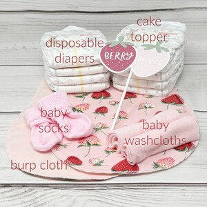 May include: A pink and white baby shower gift set with a cake topper that says "Berry Sweet". The set includes disposable diapers, baby socks, washcloths, and a burp cloth.