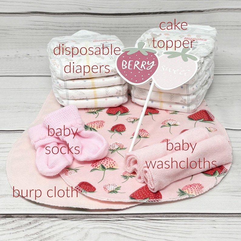May include: A pink and white baby shower gift set with a cake topper that says "Berry Sweet". The set includes disposable diapers, baby socks, washcloths, and a burp cloth.