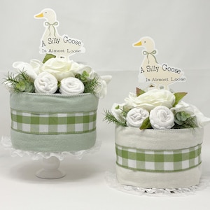 May include: Two decorative baby shower cakes. One is light green with a white and green checkered ribbon, and the other is white with a green and white checkered ribbon. Both have white flowers, rolled towels, and a sign that says "A Silly Goose Is Almost Loose".