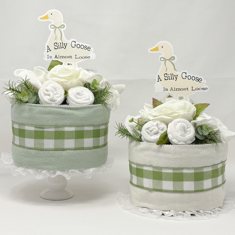 May include: Two decorative baby shower cakes. One is light green with a white and green checkered ribbon, and the other is white with a green and white checkered ribbon. Both have white flowers, rolled towels, and a sign that says "A Silly Goose Is Almost Loose".
