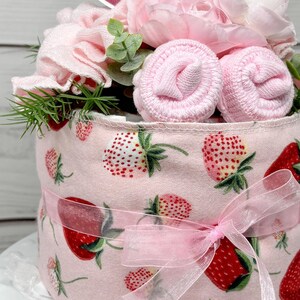 May include: A pink and white diaper cake decorated with pink flowers, green leaves, and a pink ribbon. The cake is made of diapers, baby socks, and a washcloth. The cake is decorated with a strawberry pattern.