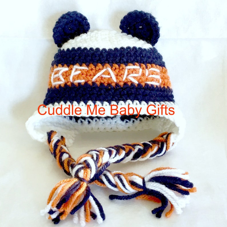 May include: White, orange, and navy blue crocheted baby hat with pom poms on top and a braided tassel. The hat has the word "BEARS" embroidered on the front.