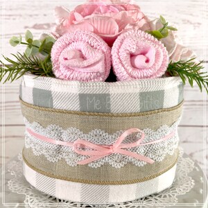 May include: A baby shower gift basket with a gray and white checkered base, wrapped with burlap ribbon and lace. The basket is decorated with pink rolled towels and pink flowers.