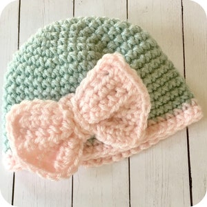 Peut inclure: Un bonnet b&eacute;b&eacute; crochet&eacute; de couleur vert clair avec un n&oelig;ud crochet&eacute; rose sur le dessus.