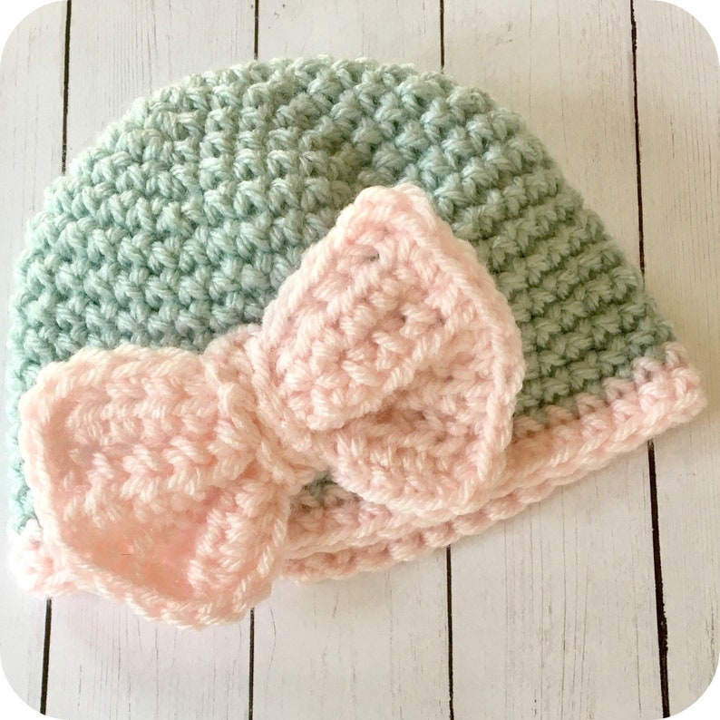 Peut inclure: Un bonnet b&eacute;b&eacute; crochet&eacute; de couleur vert clair avec un n&oelig;ud crochet&eacute; rose sur le dessus.