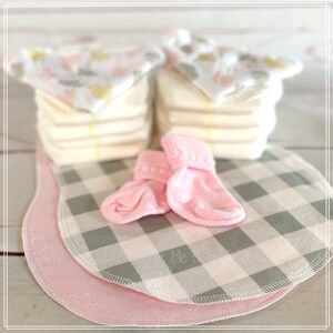 May include: A set of baby essentials including a stack of white cloth diapers, a pair of pink baby socks, and two gray and white checkered burp cloths with pink trim.