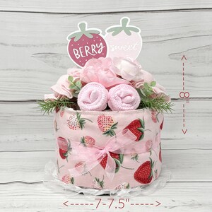 May include: A pink and white diaper cake decorated with pink flowers, green leaves, and two strawberry-shaped signs that say "Berry sweet". The cake is 7.5 inches in diameter and 8 inches tall.